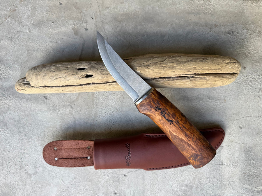 Roselli UHC Hunting Knife RW200 with dark stained handle and dark leather sheath, resting on driftwood