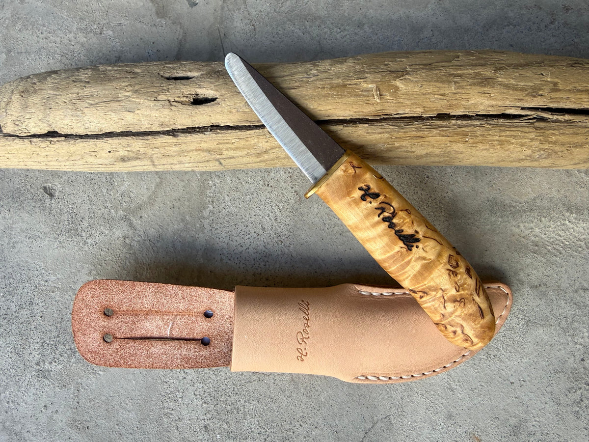 Roselli Little Carpenter R140 with rounded tip and brass guard, beside its leather sheath on driftwood